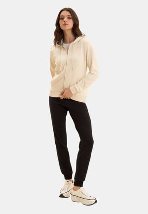 Donna in piedi che indossa una felpa con cappuccio crema con zip e fodera leopardata, pantaloni jogger neri e sneakers platform crema.