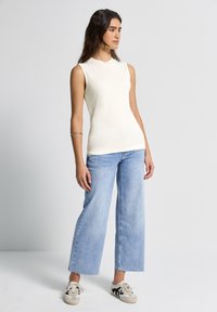 Top sin mangas de color crema con escote redondo, combinado con jeans de pierna ancha de color azul claro y zapatillas casuales. Pulsera plateada en el brazo izquierdo.