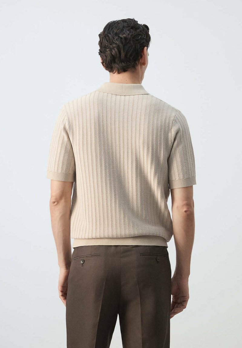 Homme portant un polo en maille beige à manches courtes et un pantalon marron foncé, debout, dos tourné, sur un fond clair uni.