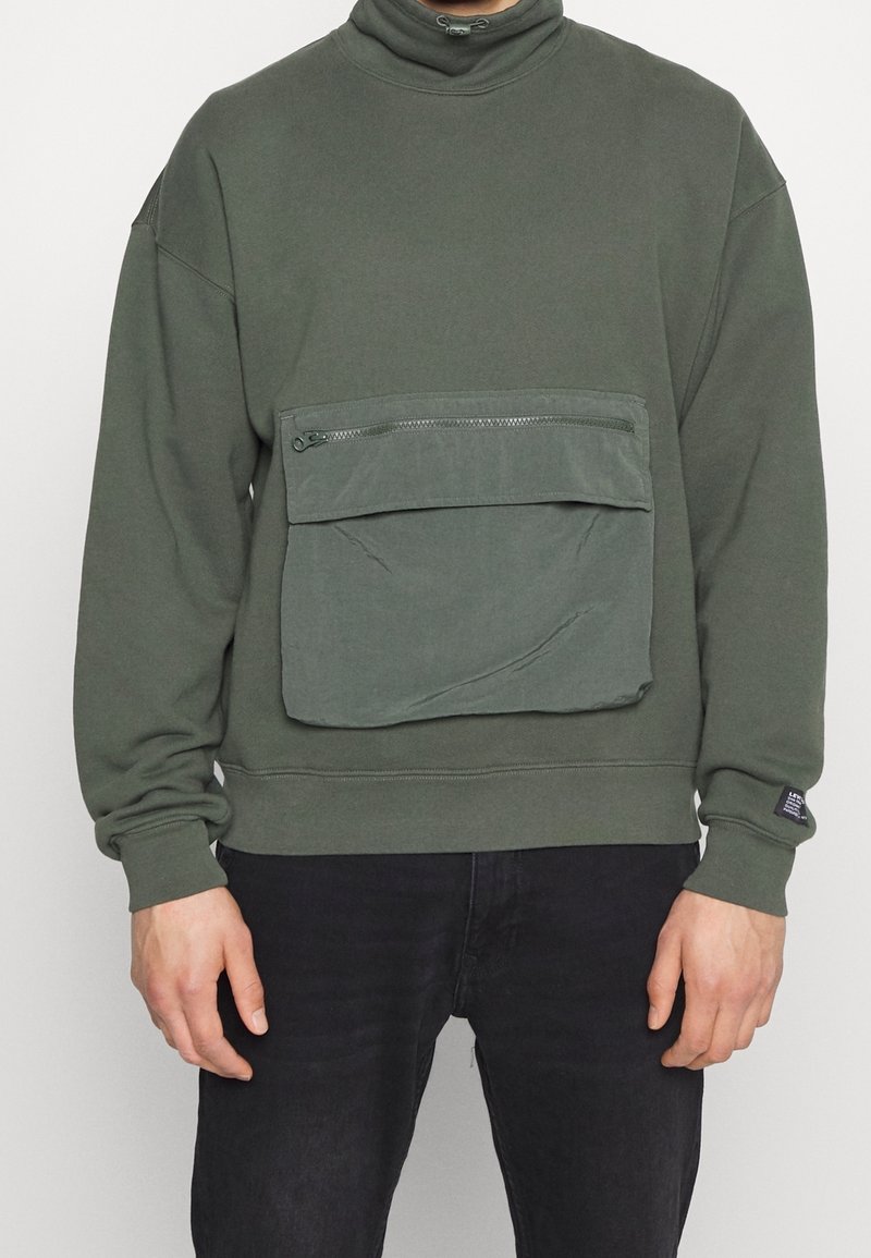 Sudadera verde oliva con cuello alto, con un gran bolsillo delantero con cierre de cremallera. Fabricada con material suave y puños acanalados.