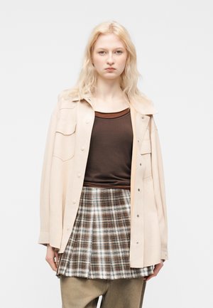 Blonde Frau trägt ein cremefarbenes Hemd über einem braunen Oberteil, karierter Rock und lockere beige Hose, steht vor einem schlichten weißen Hintergrund.