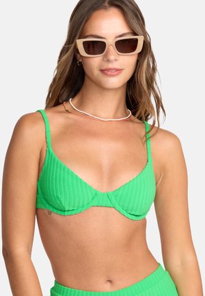 Groene geribbelde bikinitop met dunne bandjes, met een draai-detail in het midden. Model draagt beige zonnebril en gelaagde kralenkettingen.