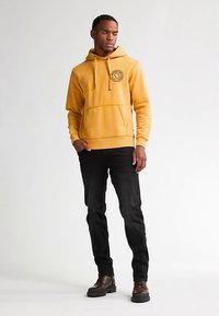 Žlutá mikina s kapucí, přední kapsou a logem, ve spojení s černými džínami a hnědými botami, stojící na světlém pozadí.
