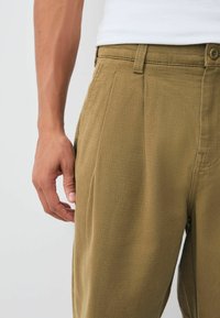 Khaki pants made of textured fabric, featuring a pleated front, slanted pockets, and a button fly, with a relaxed fit and straight leg design.