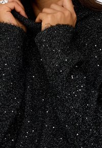 Des mains tirent le col d'un pull noir en maille avec de petites sequins brillants sur tout le tissu.