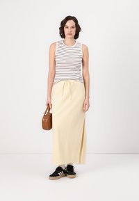 Haut sans manches rayé, jupe maxi crème-jaune avec fente sur le côté, baskets noires et sac à main marron, sur fond uni.