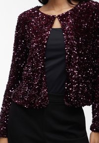 Bordinės spalvos blizgi liemenė su apvaliu iškirpimu ir atvira priekyje, turi ilgus rankovus ir aptemptą siluetą.