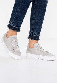 Jasnoszare płócienne trampki z białymi gumowymi podeszwami, wyposażone w sznurowany design oraz detal z logo na języku. Noszone z ciemnymi dżinsami.
