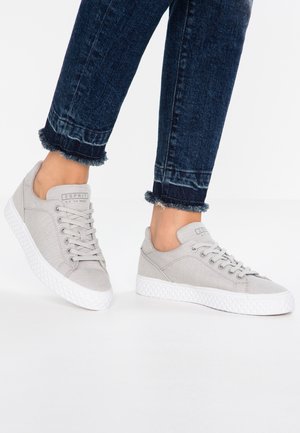 Lichtgrijze canvas sneakers met witte rubberen zolen, met een vetersluiting en merkdetail op de tong. Draagt over donkere jeans.