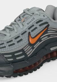 Grijze en zwarte Nike Max Air sneaker met oranje swoosh-logo en zichtbare luchtkussenzool, met getextureerd mesh en veters.