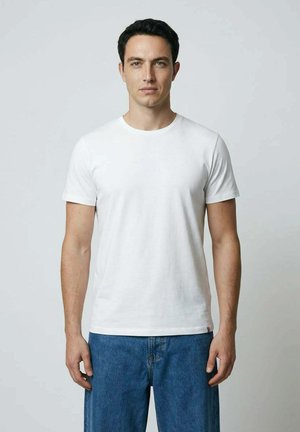 Junger Mann mit kurzem dunklem Haar, der ein schlichtes weißes T-Shirt und blaue Jeans trägt und vor einem hellgrauen Hintergrund steht.