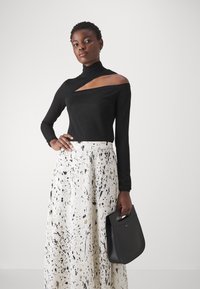 Black long-sleeve top with an asymmetrical cutout, paired with a white skirt featuring black and gold splatter patterns; holding a black handbag.