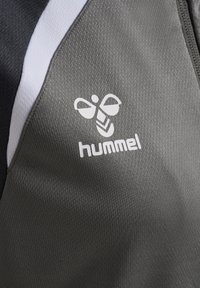 Primer plano de tela deportiva gris con logo blanco de Hummel que presenta una abeja estilizada en la zona del pecho.