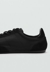 Sneaker nera con tacco in suede texturizzato, parte superiore in materiale sintetico liscio e design con lacci. I dettagli di cucitura sottili aggiungono interesse visivo.