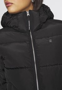 Femme portant une doudoune noire G-Star Raw avec un col montant et une fermeture éclair argentée, accessoirisée de boucles d'oreilles créoles argentées.