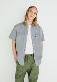 Short-sleeve denim shirt in light blue with two chest pockets and snap buttons, paired with olive green drawstring trousers.