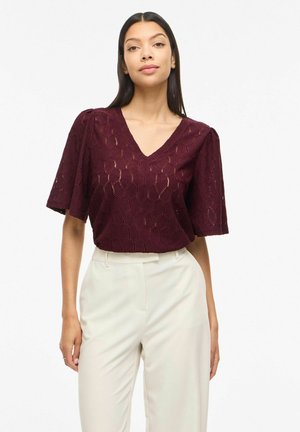 Sieviete tērpusies tumši ķiršu krāsas īsām piedurknēm ar fakturētu V-veida kakla izgriezumu un augsta vidukļa krēmkrāsas biksēs, stāvot pret baltu fonu.