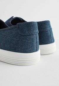 Denim slip-on sko med en mørkeblå tekstur, hvide gummisåler og minimalistisk design med en afrundet bagende og stikninger.