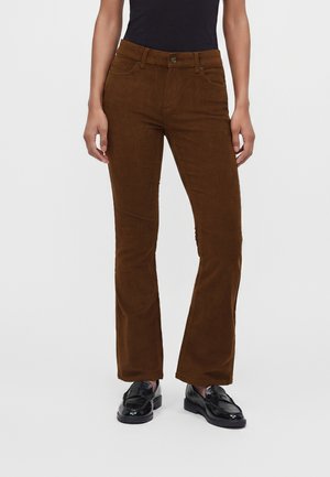 Femme portant un pantalon flare en velours côtelé marron, un haut noir et des mocassins en cuir noirs, debout devant un fond clair uni.