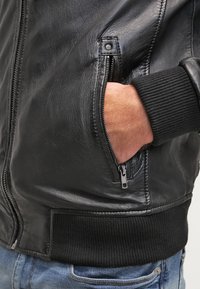Giacca bomber in pelle nera con polsini a coste, tasche laterali e dettaglio con tasca con zip, che mette in evidenza una texture liscia e un design classico.