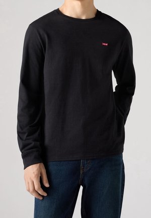 Homme portant un t-shirt noir uni à manches longues Levi's avec un petit logo rouge sur la poitrine et un jean bleu foncé sur fond neutre.