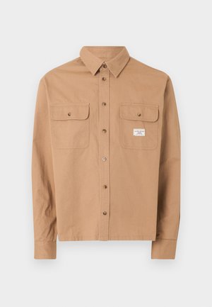 Hellbraunes langärmeliges Hemd mit durchgehendem Knopfverschluss, zwei geknöpften Brusttaschen und einem kleinen weißen rechteckigen Etikett auf der rechten Tasche.