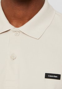 Gros plan sur un polo beige avec deux boutons et un patch logo rectangulaire noir Calvin Klein sur la poitrine.
