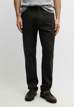 Homme portant un jean gris foncé à coupe droite, une chemise grise claire rentrée à l'intérieur et des chaussures en cuir marron à lacets, debout devant un fond uni.