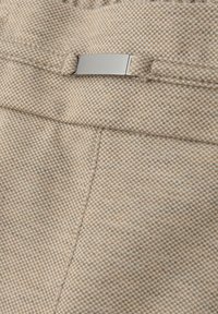 Tissu texturé beige avec un motif à carreaux et une étiquette métallique lisse. Présente des détails de couture et une poche avant.