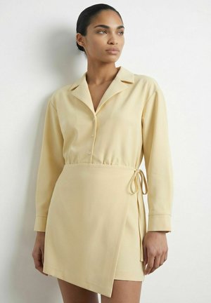 Femme portant une robe portefeuille beige à manches longues avec des boutons et un nœud sur le côté, debout devant un fond blanc uni, regardant sur le côté.