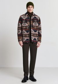 Gemusterte Wolljacke mit geometrischen Designs in erdigen Tönen, kombiniert mit einem dunklen Rollkragenpullover und braunen Hosen, abgerundet mit Lederstiefeln.