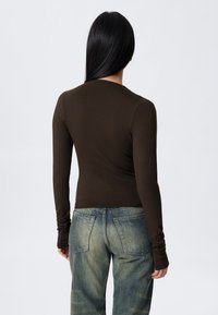 Hnědý top s dlouhým rukávem a přiléhavým střihem; má kulatý výstřih a prodloužené manžety, doplněný vybledlými modrými džínami.