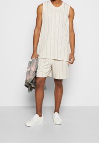 Beige ärmlös topp och short-set med teal vertikala ränder; strukturerad tyg. Vita sneakers och en flerfärgad rutig jacka.