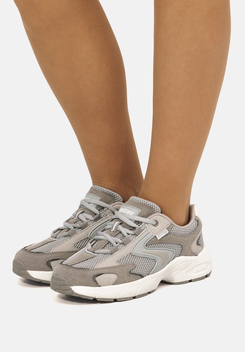 Zapatillas de malla y ante en gris y blanco con cordones, usadas sobre pies desnudos contra un fondo claro y sencillo.