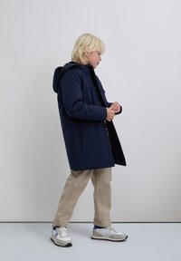 Kind met blond haar gekleed in een marineblauwe hoodie, beige broek en witte sneakers, staande tegen een effen lichte achtergrond.