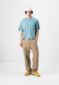 Lys blå t-skjorte med korte ermer, khaki cargo-bukser og gule joggesko. Modellen har på seg en hvit caps. Teksturert stoff med et subtilt stripemønster.
