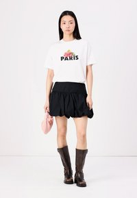Fehér póló "PARIS" felirattal és virágos grafikával, fekete fodros szoknyával és barna térdcsizmával párosítva, rózsaszín táskával a kezében.