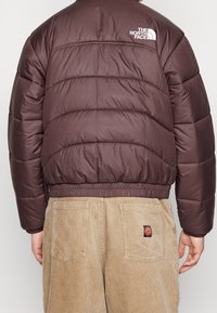 Brązowa puchowa kurtka z pikowanym wzorem i elastycznym dołem. Logo na lewym ramieniu. Połączona z beżowymi spodniami z korduroju z łatą.