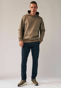 Beige Kapuzenpullover mit einer Fronttasche und Kordelzug, kombiniert mit marineblauen Slim-Fit-Hosen. Das Modell trägt beigemfarbene Sneakers mit strukturierten Akzenten.