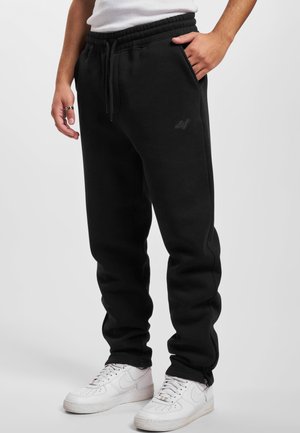 Person, die schwarze Jogginghose mit elastischem Bund und weißen Sneakers trägt, steht mit einer Hand in der Tasche vor einfarbigem Hintergrund.