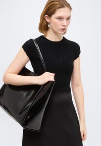 Γυναίκα που φοράει μαύρη πλεκτή μπλούζα με κοντά μανίκια και φούστα, κρατώντας μια μεγάλη, γυαλιστερή μαύρη τσάντα στον ώμο της.