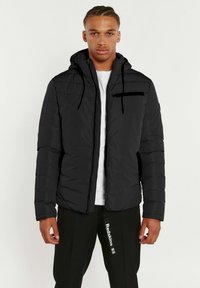 Veste matelassée noire avec capuche, poche zippée sur la poitrine, tissu texturé et cordons de serrage ajustables. Portée par-dessus une chemise blanche.