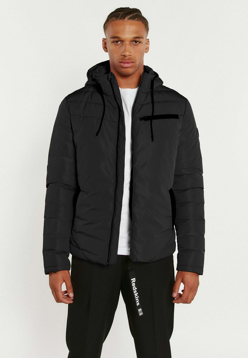 Veste matelassée noire avec capuche, poche zippée sur la poitrine, tissu texturé et cordons de serrage ajustables. Portée par-dessus une chemise blanche.