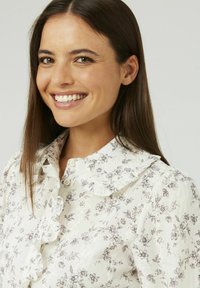 Blouse blanche avec un motif floral brun ; présente un col froncé et des boutons sur le devant. Le tissu semble léger avec une finition texturée.