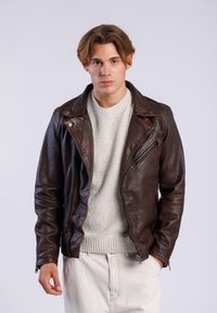 Veste de moto en cuir marron avec fermeture à glissière asymétrique, texture mate et détails en métal, portée sur un pull en maille gris clair.