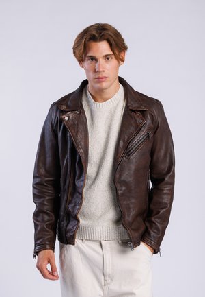 Chaqueta de cuero marrón tipo biker con cierre de cremallera asimétrico, textura mate y detalles de metal, llevada sobre un suéter de punto gris claro.