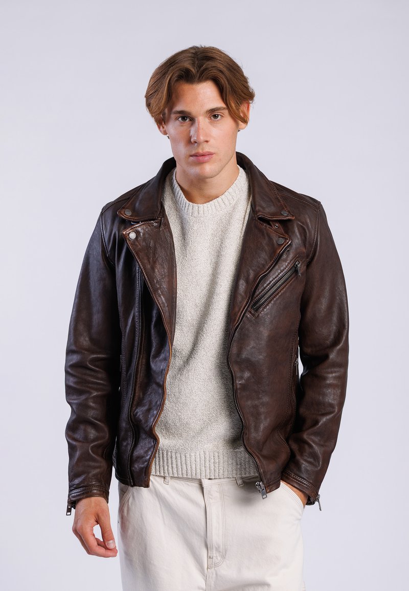 Veste de moto en cuir marron avec fermeture à glissière asymétrique, texture mate et détails en métal, portée sur un pull en maille gris clair.