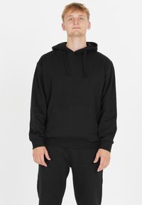 Nuori mies seisoo, päällään yksinkertainen musta huppari etutaskulla ja matching mustat housut valkoista taustaa vasten.