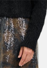 Schwarzer, flauschiger Pullover mit langen Ärmeln über einem glitzernden Rock, der mit einem verlaufenden Muster aus Silber- und Goldtönen verziert ist.