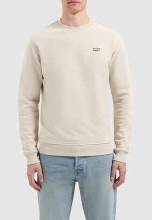 Beige katoenen sweatshirt met een ronde hals, geribbelde manchetten en zoom, voorzien van een klein "PURE PATH" logo op de linkerborst. In combinatie met lichtblauwe jeans.